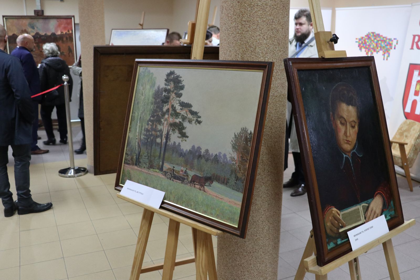 Wystawa dwóch obrazów. Obraz z lewej strony prezentuje trzy osoby siedzące w powozie ciągniętym przez konia na tle lasów. Na obrazie z prawej strony namalowany został portret kobiety zamyślonej notującej piórem na księdze.