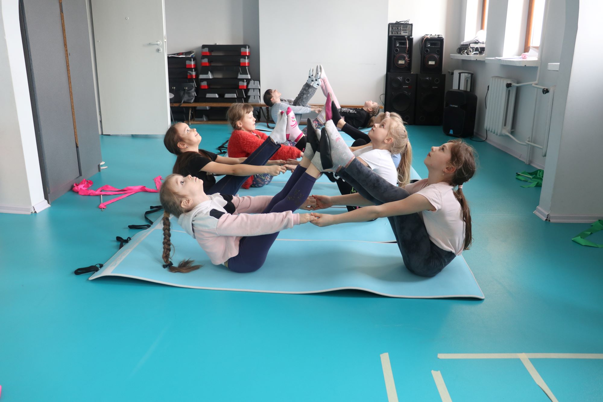 Grupa dziewczynek z zajęć artystyczno ruchowych ćwiczy na matach rozciąganie mięśni. Dziewczynki łączą się stopami łapią się za ręce i odchylają głowy do tyłu.